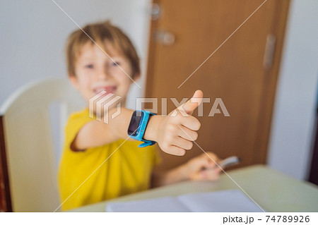 Little boy sitting at the table and looking smart watch. Smart watch for baby safety. The child makes school lessons, listening to music, calling friends 74789926