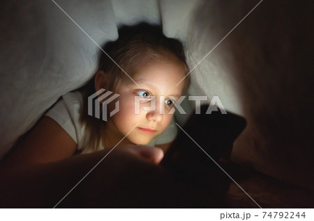 Child watching video smartphone under the blanket on the bed at night when light flashes bounce off the screen, children use addiction games and cartoon concept 74792244