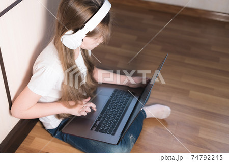 A little girl in white headphones with a laptop on her lap sits on the floor. look with a serious face in your laptop 74792245