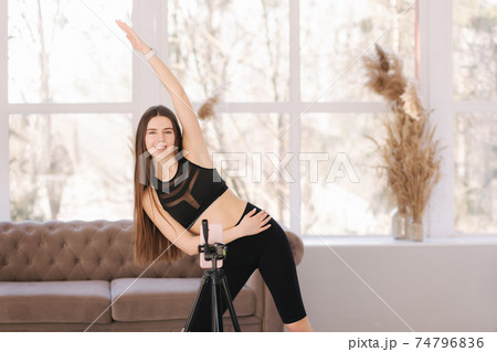 Young athletic woman blogger in balck sportswear shoots video on phone as she does exercises at home in living room. Sport and recreation concept. Fitness vlogger recording live tutorial video Young athletic woman blogger in balck sportswear shoots video on phone as she does exercises at home in living room. Sport and recreation concept. Fitness vlogger recording live tutorial video 74796836