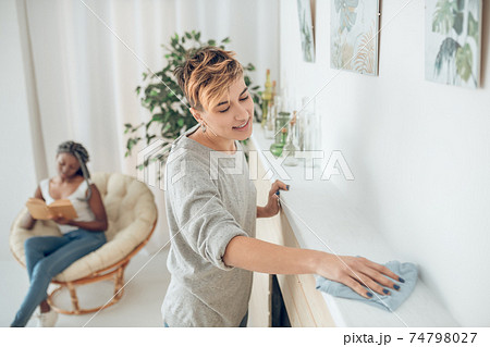 Fair-haired girl cleaning the room, her girlfriend sitting in the chair Fair-haired girl cleaning the room, her girlfriend sitting in the chair 74798027