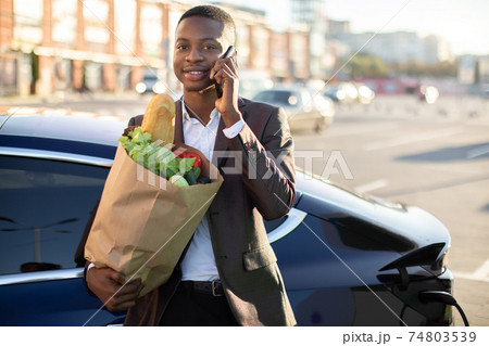 Smiling young afro american businessman, holding paper bag with food, talking on mobile phone and looking at camera, while his electric car is charging at city EV charging station Smiling young afro american businessman, holding paper bag with food, talking on mobile phone and looking at camera, while his electric car is charging at city EV charging station 74803539