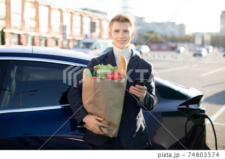 Front view of young happy pleasant guy in trendy formal wear, feeling relaxed after shopping, using phone while refueling his modern luxury electric car at the city station Front view of young happy pleasant guy in trendy formal wear, feeling relaxed after shopping, using phone while refueling his modern luxury electric car at the city station 74803574