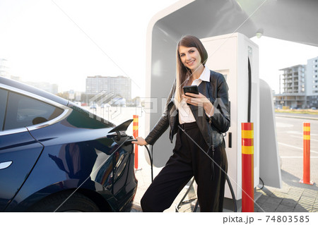 smiling young adult woman paying for charging electric car, holding smartphone in hand and looking at camera. Happy girl standing at public charging station near electric automobile smiling young adult woman paying for charging electric car, holding smartphone in hand and looking at camera. Happy girl standing at public charging station near electric automobile 74803585