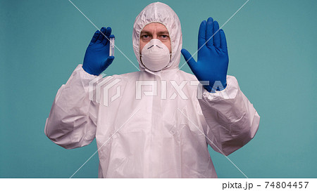 A male doctor in protective overalls and a mask holds an ampoule with a vaccine in his hands.  74804457