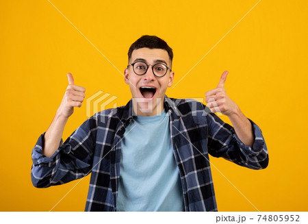 Funky young guy in glasses showing thumbs up with both hands, approving of something on orange studio background Funky young guy in glasses showing thumbs up with both hands, approving of something on orange studio background 74805952