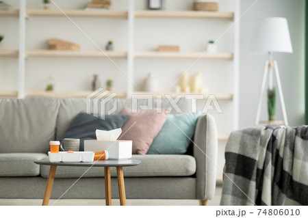 Napkins in box on table, cup of hot drink and medications in jars, gray sofa with colored pillows and blanket Napkins in box on table, cup of hot drink and medications in jars, gray sofa with colored pillows and blanket 74806010