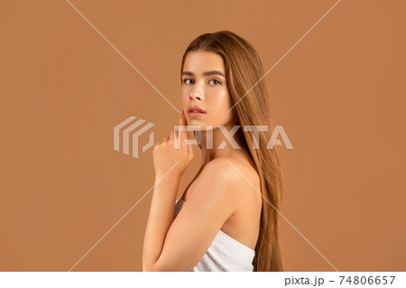 Millennial woman with long straight hair touching her face, having soft healthy skin and nude makeup on brown background 74806657