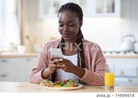 Smiling Black Woman Texting On Smartphone While Having Breakfast In Kitchen 74806658