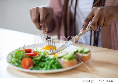 Unrecognizable african woman eating tasty breakfast in kitchen, using fork and knife Unrecognizable african woman eating tasty breakfast in kitchen, using fork and knife 74806661