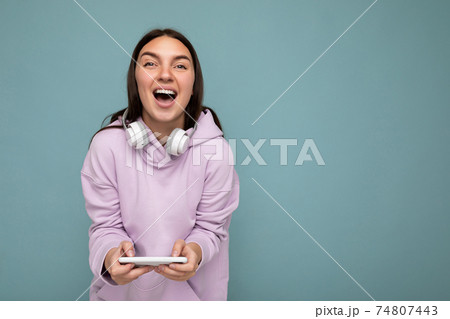 Pretty positive laughing young brunette woman wearing purple hoodie isolated on blue background Pretty positive laughing young brunette woman wearing purple hoodie isolated on blue background 74807443