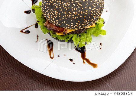 Black burger on a white plate. Beef patty burger, cheddar cheese, lettuce, bacon, tomato and balsamic sauce. The bun is made with the addition of ink cuttlefish. Top View. 74808317