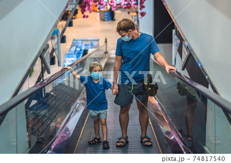 Front view boy, father going up escalator in super market wear face protect mask, COVID coronavirus pandemic lockdown quarantine. Dad hold child hand, look at kid. New life reality, family shopping Front view boy, father going up escalator in super market wear face protect mask, COVID coronavirus pandemic lockdown quarantine. Dad hold child hand, look at kid. New life reality, family shopping 74817540
