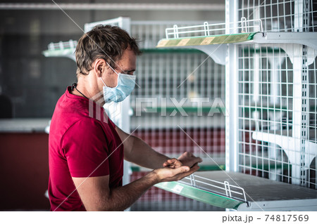 Man wear blue protective mask look on stripped bare shelves in empty shop store, COVID pandemic social panic. Hand gesture show stress question, afraid of isolation lockdown crisis problems. Real skin 74817569
