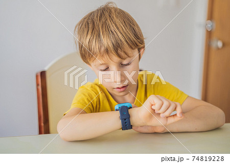 Little boy sitting at the table and looking smart watch. Smart watch for baby safety. The child makes school lessons, listening to music, calling friends Little boy sitting at the table and looking smart watch. Smart watch for baby safety. The child makes school lessons, listening to music, calling friends 74819228