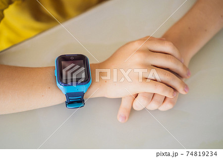 Little boy sitting at the table and looking smart watch. Smart watch for baby safety. The child makes school lessons, listening to music, calling friends Little boy sitting at the table and looking smart watch. Smart watch for baby safety. The child makes school lessons, listening to music, calling friends 74819234