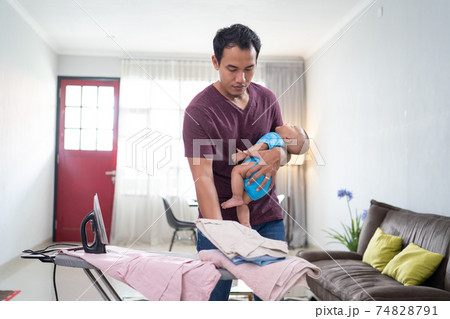 father ironing his clothes while holding his infant baby on his hand 74828791