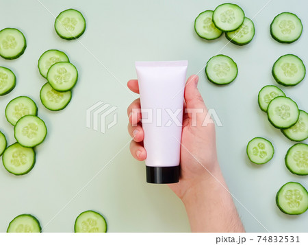 Male hand holds plastic unbranded pink cream tube with black cap. Bottle for branding and label and fresh sliced cucumber on green background. Natural organic spa cosmetics. Skincare, advertising Male hand holds plastic unbranded pink cream tube with black cap. Bottle for branding and label and fresh sliced cucumber on green background. Natural organic spa cosmetics. Skincare, advertising 74832531