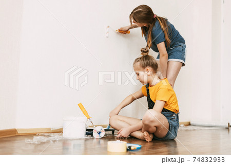 A girl with her sister in bright blue and yellow clothes helps to paint the walls in her room white A girl with her sister in bright blue and yellow clothes helps to paint the walls in her room white 74832933