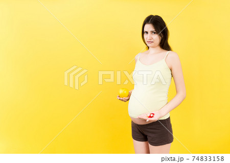 pregnant woman making a choice between an apple and a pile of pills at colorful background with copy space. Taking vitamins during pregnancy concept pregnant woman making a choice between an apple and a pile of pills at colorful background with copy space. Taking vitamins during pregnancy concept 74833158