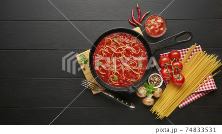 Cooking spaghetti with tomato sauce in cast iron pan served with red chili pepper, fresh basil, cherry-tomatoes and spices over black rustic wooden background. 74833531