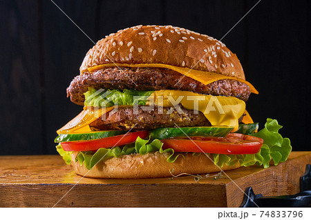 Closeup of home made beef burger with lettuce and tomatoes served on wooden cutting board. Dark background Closeup of home made beef burger with lettuce and tomatoes served on wooden cutting board. Dark background 74833796