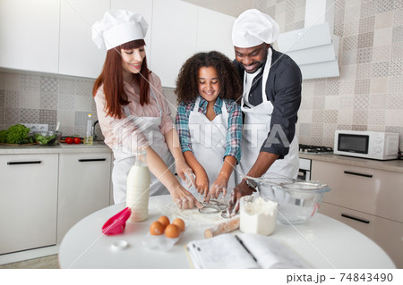 Multiethnic parents and their mixed race curly daughter squeezing round shapes out of the dough with cookies cutters 74843490