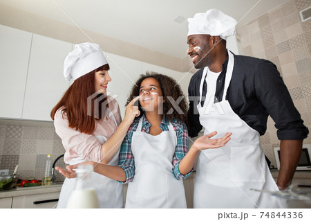Close up of funny crazy multiracial family, father, mother and teen girl, preparing pastry in the kitchen, fooling and playing together with flour and enjoy pastime. Close up of funny crazy multiracial family, father, mother and teen girl, preparing pastry in the kitchen, fooling and playing together with flour and enjoy pastime. 74844336