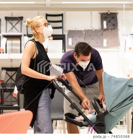 Young couple with newborn in stroller shopping at retail furniture and home accessories store wearing protective medical face mask to prevent spreading of corona virus when shops reopen. 74850388