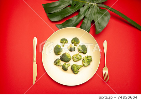 Plate of fresh raw broccoli with cutlery placed on red table near Monstera leaf Plate of fresh raw broccoli with cutlery placed on red table near Monstera leaf 74850604