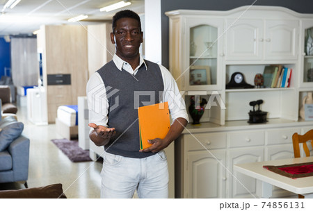 Portrait of smiling salesman who is offering modern furniture and standing with folder 74856131
