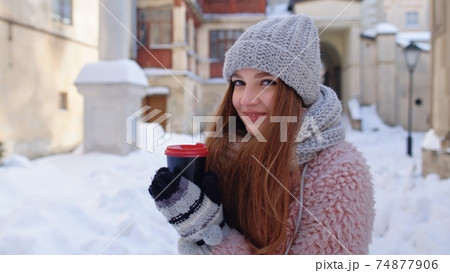 Stylish woman traveler with hot drink in cup looking around through city street during vacations 74877906