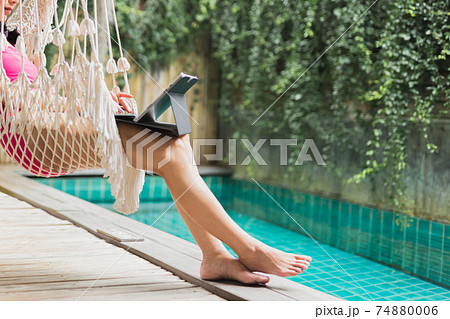 Woman in swimming suit sit in hammock working on laptop by swimming pool. 74880006