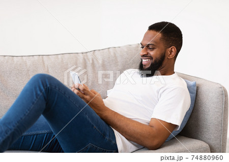 Black Guy Using Smartphone Texting Lying On Couch At Home 74880960