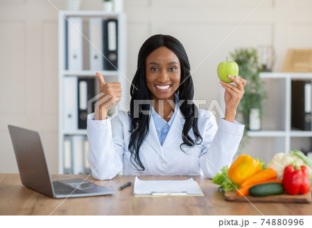 Millennial black dietitian holding apple and showing thumb up gesture at desk in medical office. Healthy eating concept 74880996