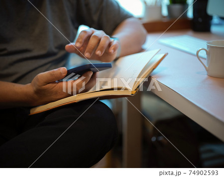 Male hand using smartphone while reading a book on computer desk in home office Male hand using smartphone while reading a book on computer desk in home office 74902593