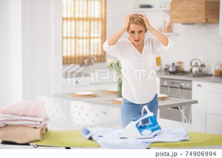 Pretty blonde woman standing near ironing table and looking desperate Pretty blonde woman standing near ironing table and looking desperate 74906994