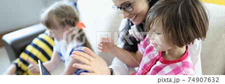 Woman is sitting on the couch, girl is sitting on her lap, they are looking at laptop next to children are playing. Woman is sitting on the couch, girl is sitting on her lap, they are looking at laptop next to children are playing. 74907632