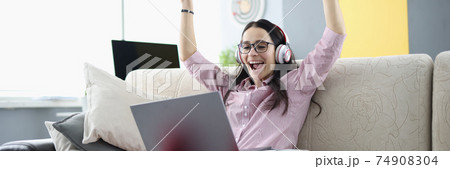 Rejoicing woman is sitting on sofa with in headphones with laptop. 74908304