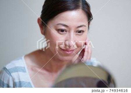 鏡を見て頬に手を当てる笑顔の中年女性 74934616