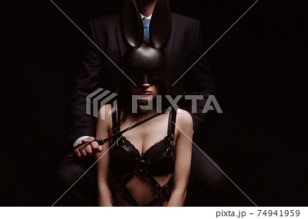 dominant man with a Flogger whip and a submissive girl in a mask and leather belt 74941959