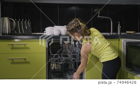 Smart girl learning to use dishwasher. Stylish modern Built In Kitchen Appliances in green black. Child is putting dirty dishes. 74946426