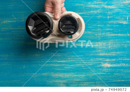 Two disposable paper cups of coffee and a cup holder in a male hand on a blue wooden background. Two disposable paper cups of coffee and a cup holder in a male hand on a blue wooden background. 74949072