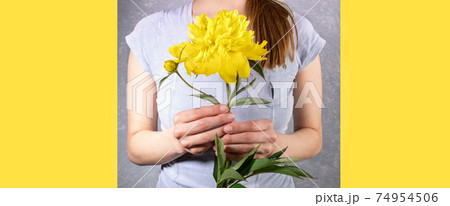Young woman faceless in grey T-shirt holding yellow peony close up on grey and yellow Illumination backgrounds. Festive banner in trendy colors of 2021 for Mother's day or Women holiday. Copy space. 74954506