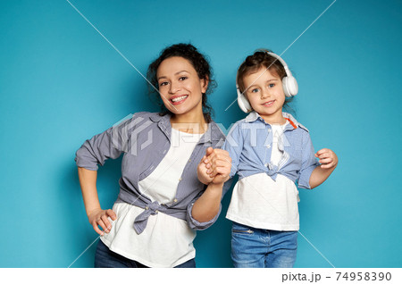 Attractive woman posing in front of the camera with a toothy smile on a blue background with her little daughter in headphones Attractive woman posing in front of the camera with a toothy smile on a blue background with her little daughter in headphones 74958390