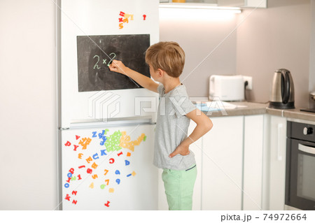 A 8-year-old schoolboy writes white chalk on a magnetic black board attached to a fridge 74972664