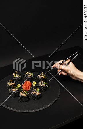 Hand with chopsticks wants to take custom sushi roll with black rice, crab meat, avocado, smoked salmon mousse, oar caviar, masago, shrimp cocktail, edible gold leaf, ginger, wasabi on black table. Hand with chopsticks wants to take custom sushi roll with black rice, crab meat, avocado, smoked salmon mousse, oar caviar, masago, shrimp cocktail, edible gold leaf, ginger, wasabi on black table. 74976135