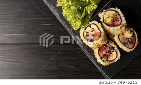 Custom sushi roll in tempura with nori, fresh salmon, tuna, avocado, masago caviar, drizzled with pineapple sauce with salad pouring as decoration on a black plate on a wooden table and background. Custom sushi roll in tempura with nori, fresh salmon, tuna, avocado, masago caviar, drizzled with pineapple sauce with salad pouring as decoration on a black plate on a wooden table and background. 74976215