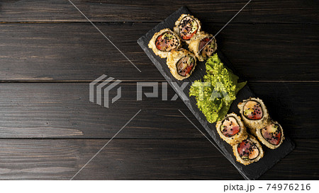 Custom sushi roll in tempura with nori, fresh salmon, tuna, avocado, masago caviar, drizzled with pineapple sauce with salad pouring as decoration on a black plate on a wooden table and background. Custom sushi roll in tempura with nori, fresh salmon, tuna, avocado, masago caviar, drizzled with pineapple sauce with salad pouring as decoration on a black plate on a wooden table and background. 74976216
