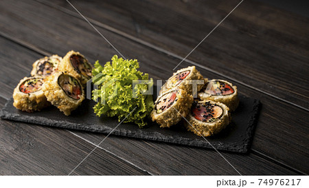 Custom sushi roll in tempura with nori, fresh salmon, tuna, avocado, masago caviar, drizzled with pineapple sauce with salad pouring as decoration on a black plate on a wooden table and background. 74976217